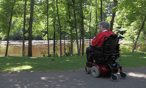 Image of a woman sitting in a power chair next to trees by a river